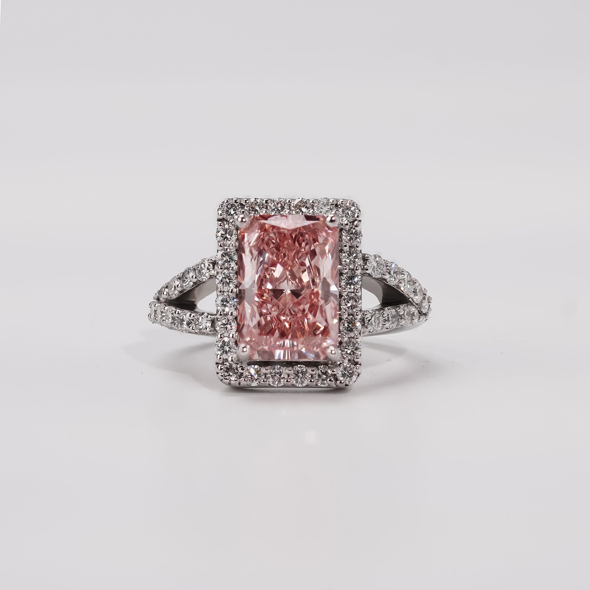 A radiant-cut pink diamond ring surrounded by a halo of small white diamonds, set on a split-shank band adorned with additional white diamonds in a white gold or platinum setting.