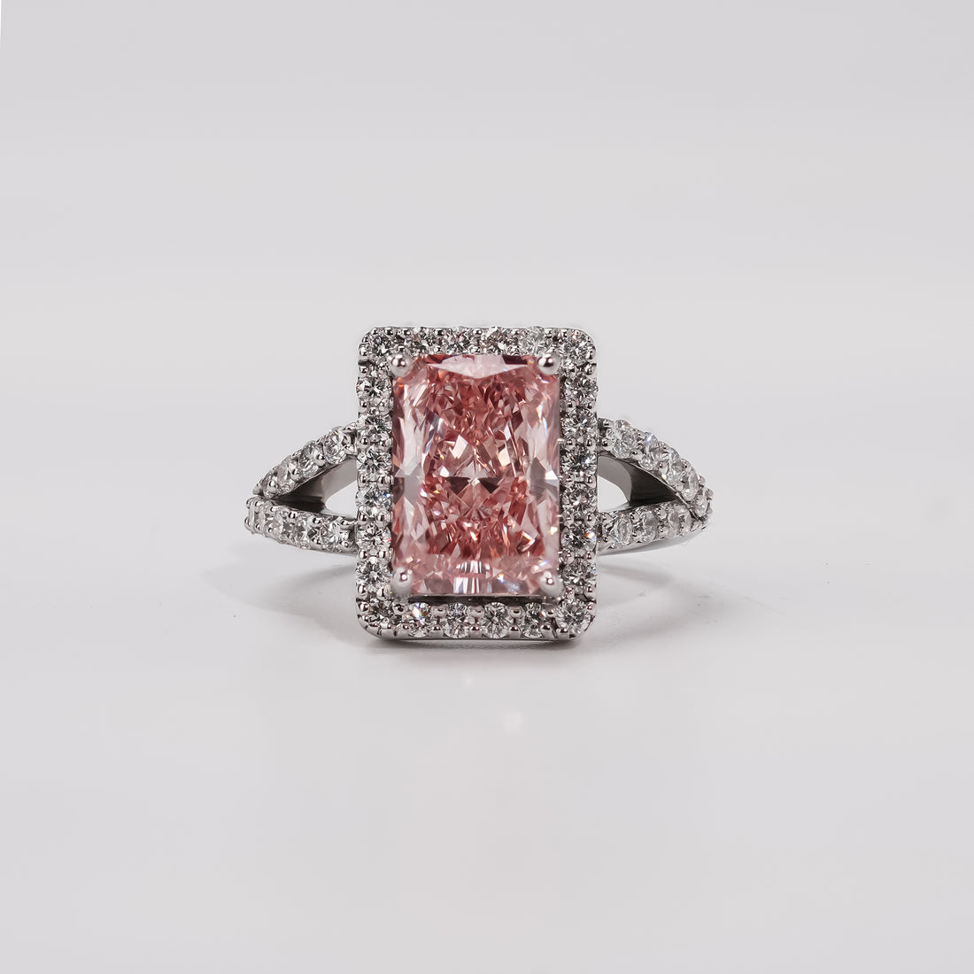 A radiant-cut pink diamond ring surrounded by a halo of small white diamonds, set on a split-shank band adorned with additional white diamonds in a white gold or platinum setting.
