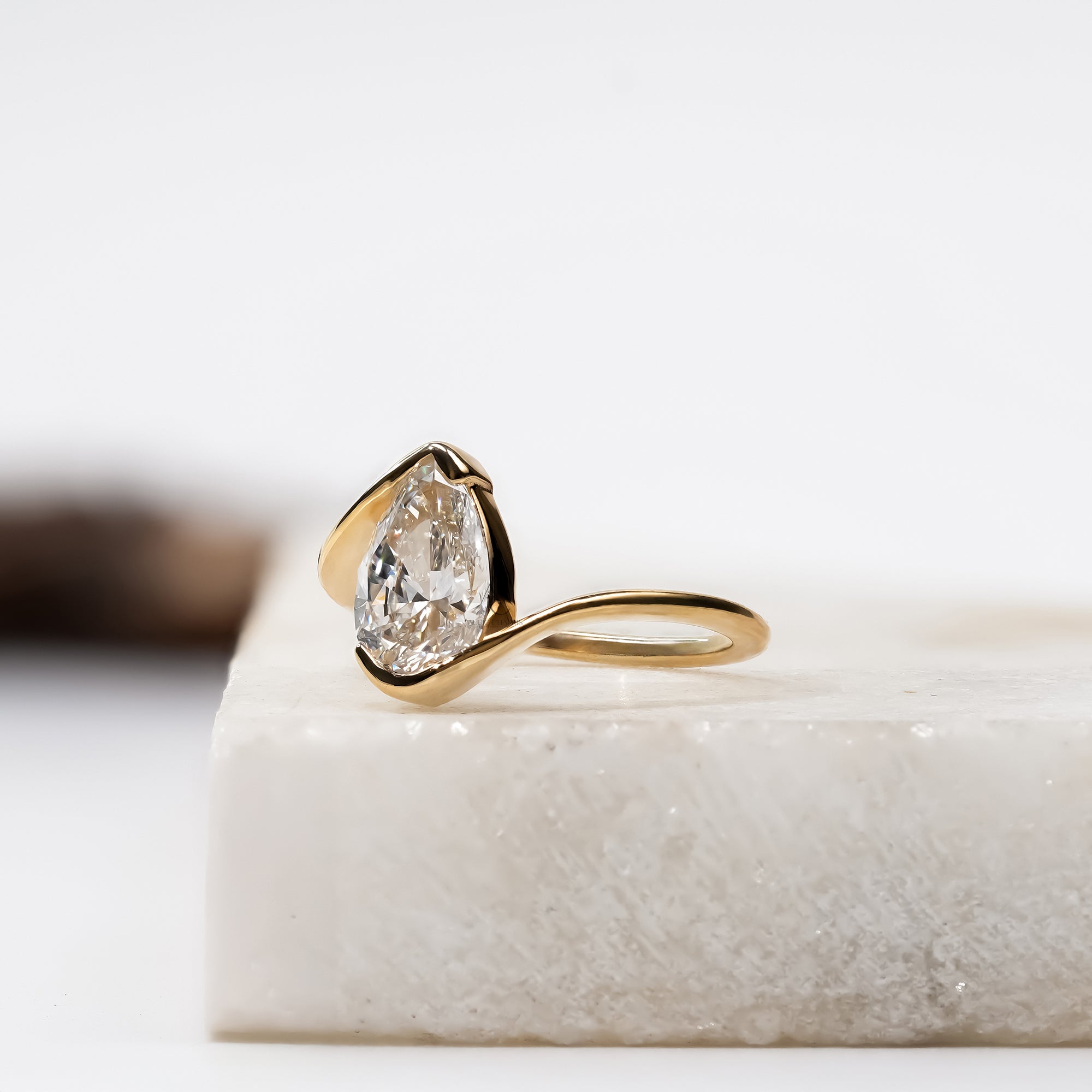A pear-shaped diamond set in a sleek, curved gold band, displayed on a light stone block in a minimalist studio setting.