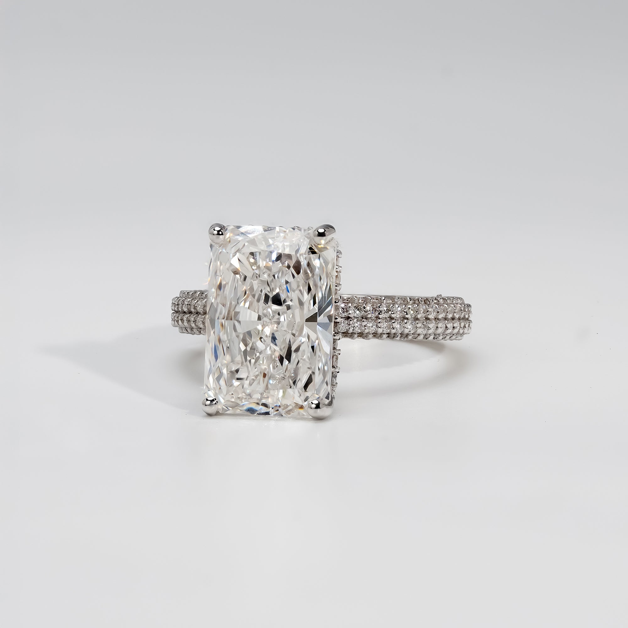 A platinum engagement ring featuring a radiant-cut diamond at the center, held by four prongs. The band is adorned with three rows of pavé-set diamonds, adding brilliance and a luxurious sparkle to the classic design.