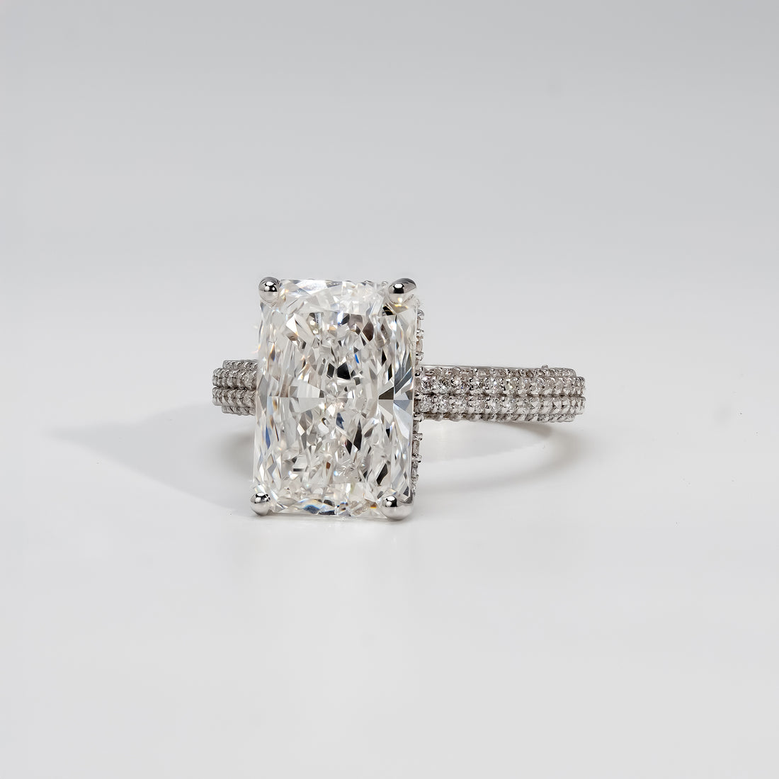A platinum engagement ring featuring a radiant-cut diamond at the center, held by four prongs. The band is adorned with three rows of pavé-set diamonds, adding brilliance and a luxurious sparkle to the classic design.