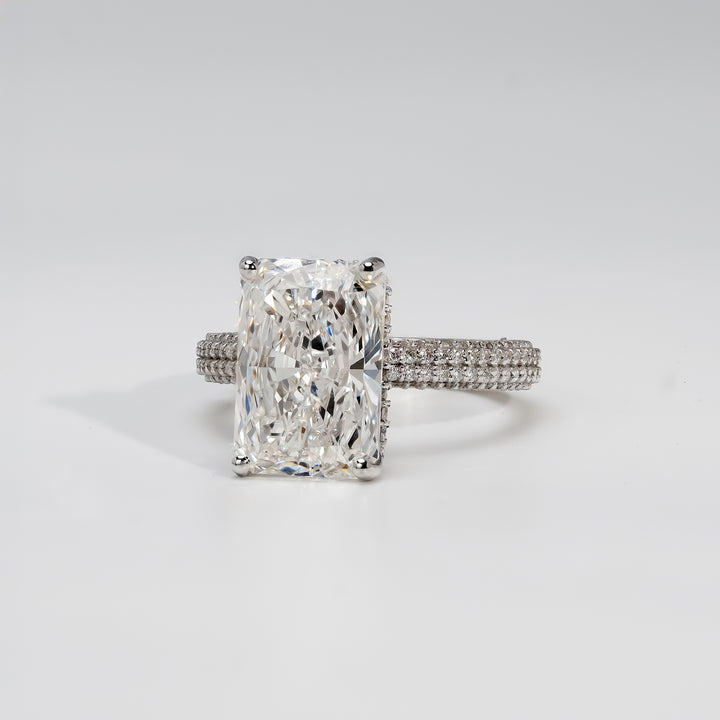 A platinum engagement ring featuring a radiant-cut diamond at the center, held by four prongs. The band is adorned with three rows of pavé-set diamonds, adding brilliance and a luxurious sparkle to the classic design.