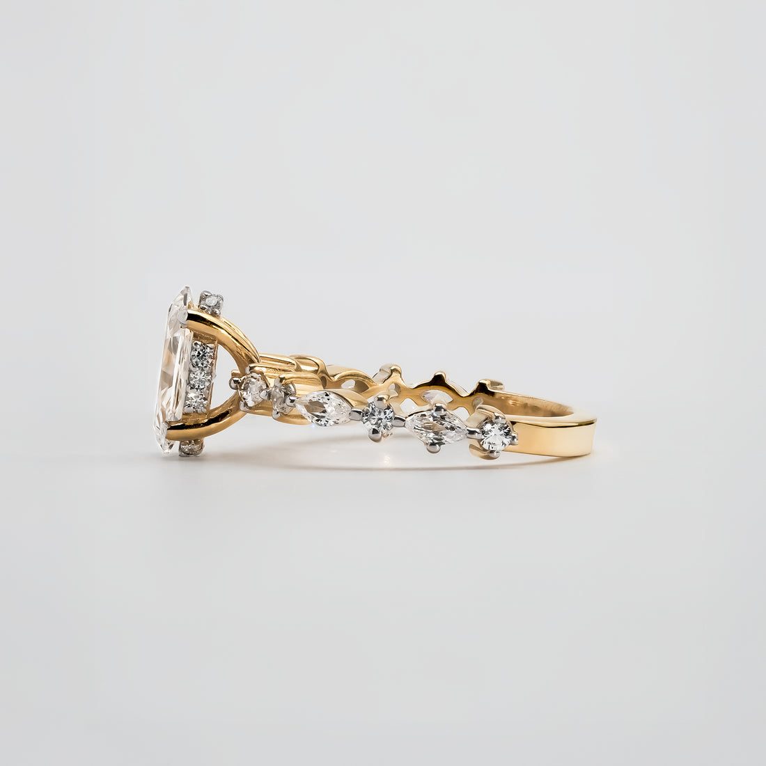 A yellow gold ring with a marquise-cut diamond centerpiece set in six prongs. The band features alternating marquise and round-cut diamonds in a woven design, adding sparkle and sophistication to the elegant setting.