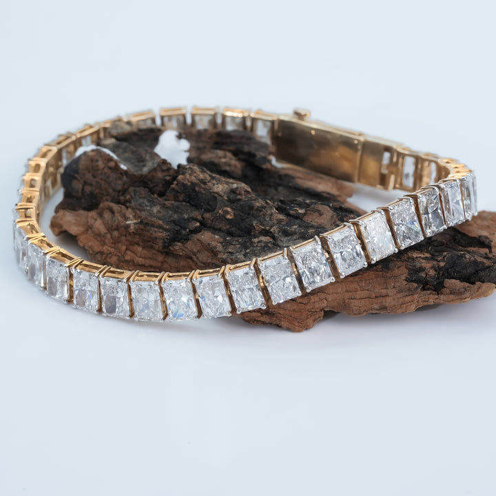 A gold bracelet featuring a continuous row of radiant-cut diamonds set in a channel-style design. The bracelet rests on a textured white surface, highlighting the brilliance of the diamonds and the warm tone of the gold setting.