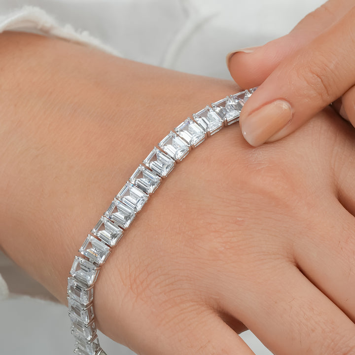 A woman’s wrist wearing a silver bracelet featuring rectangular, emerald-cut stones set in a continuous row. The bracelet shines against a white, textured sleeve, highlighting its modern and elegant design.
