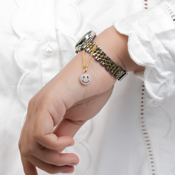 A wrist wearing a two-tone silver and gold bracelet watch. A delicate gold chain is attached to the band, featuring a small, diamond-studded smiley face charm that adds a playful sparkle to the elegant design.