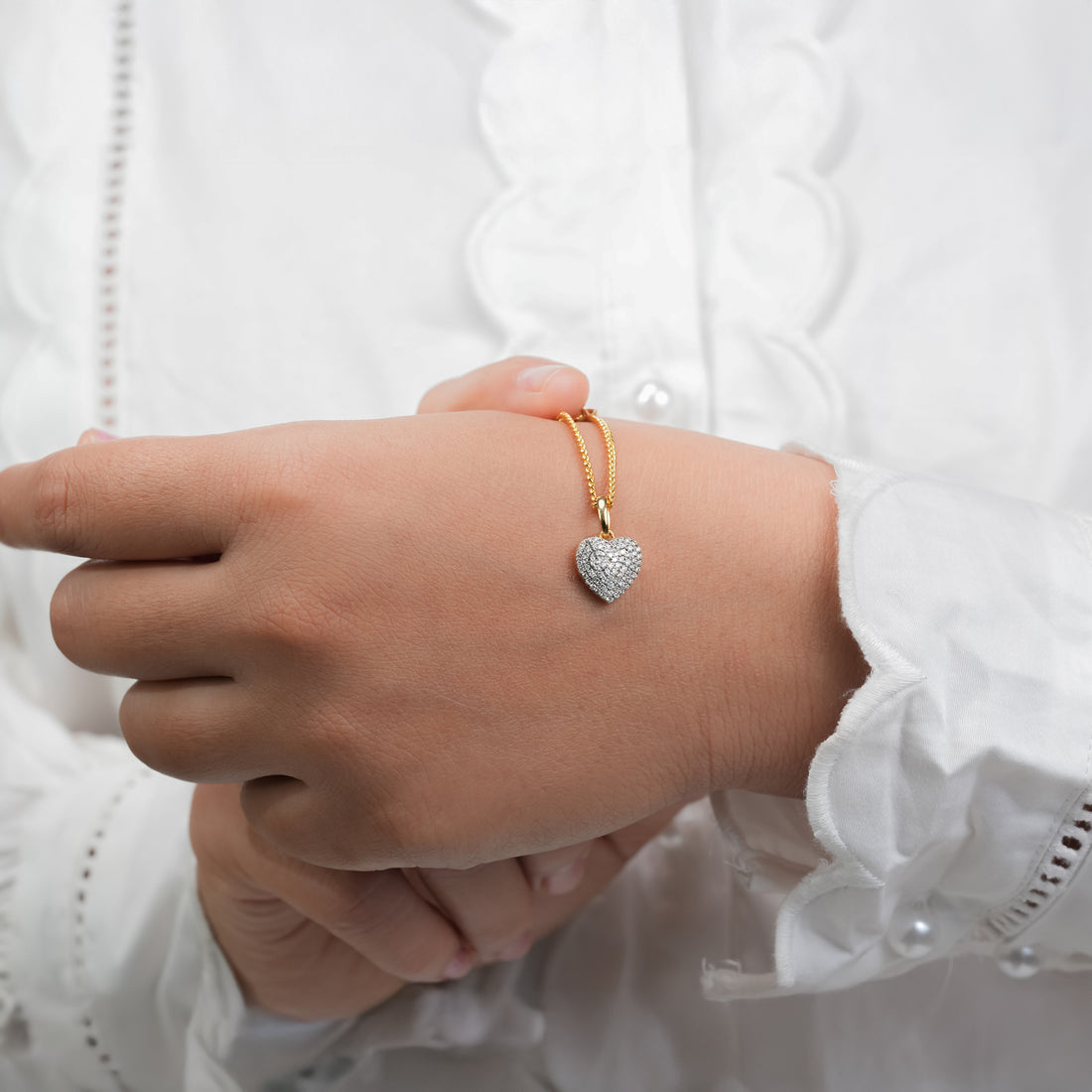 A wrist wearing a two-tone silver and gold metal watch, adorned with a gold chain and a heart-shaped diamond pendant charm. The heart charm sparkles against the white sleeve, adding an elegant and feminine touch.