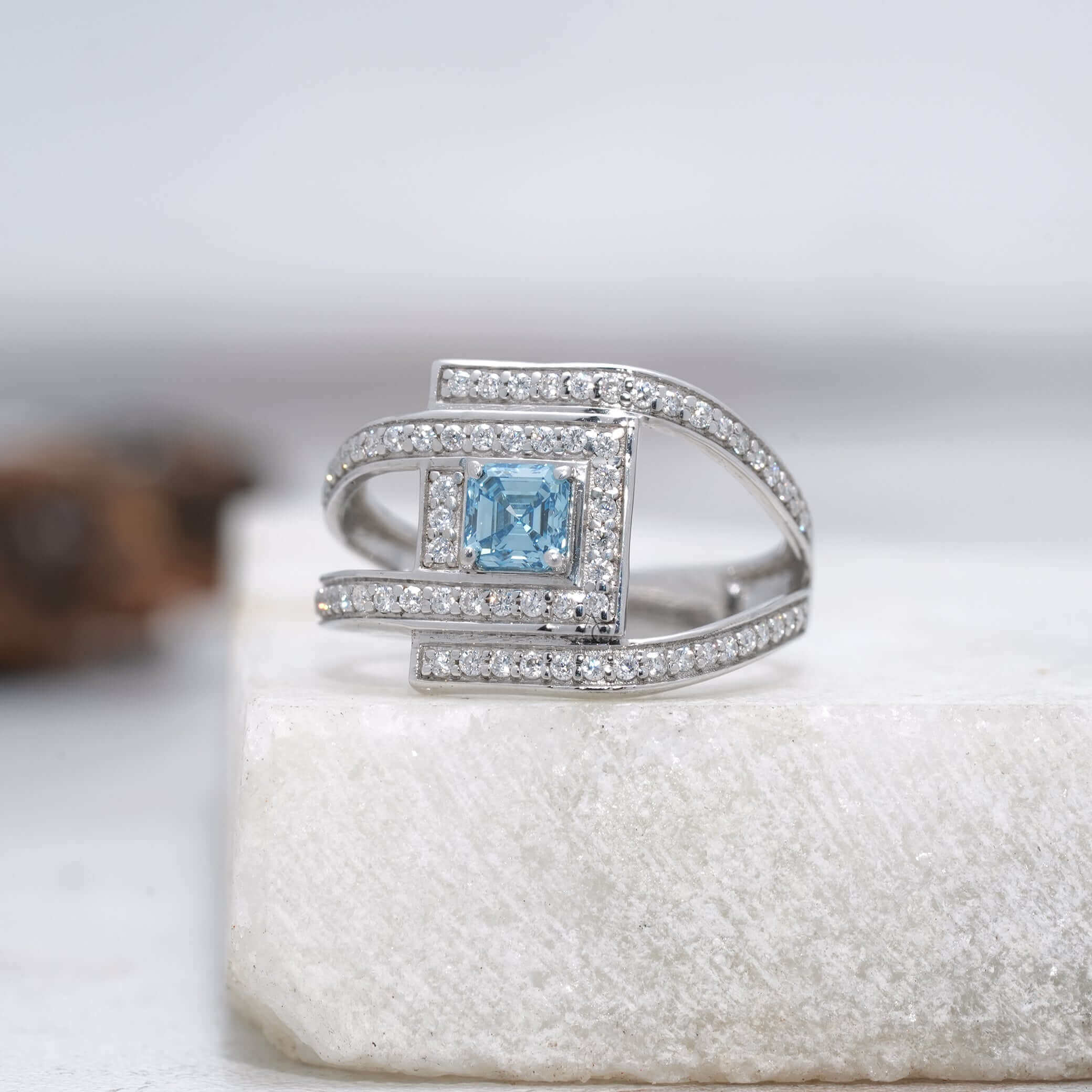 Side view of square blue gemstone ring with diamond halo on white surface.