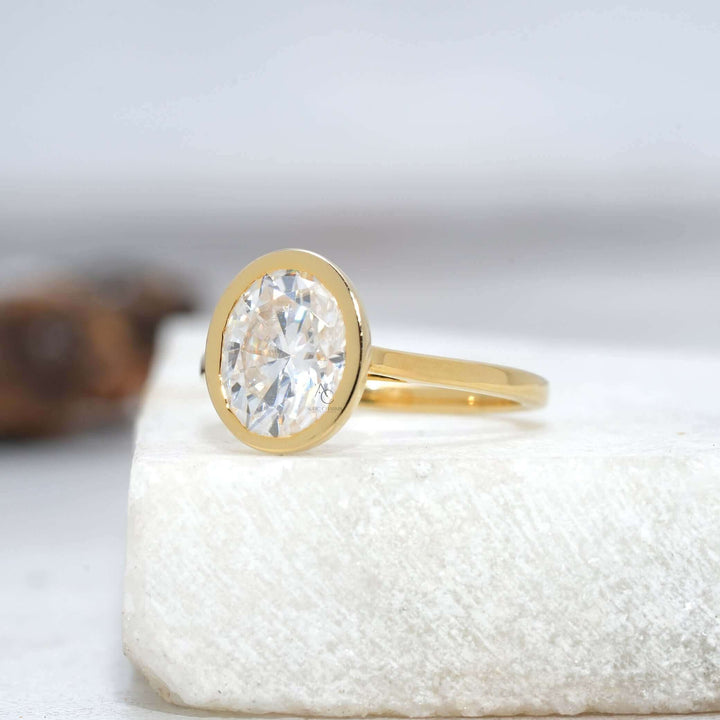 Solitaire oval diamond ring with bezel setting on reflective surface.