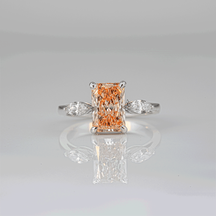 Elegant radiant-cut champagne-colored lab-grown diamond ring on a sleek white gold band with marquise side stones, captured against a reflective surface.