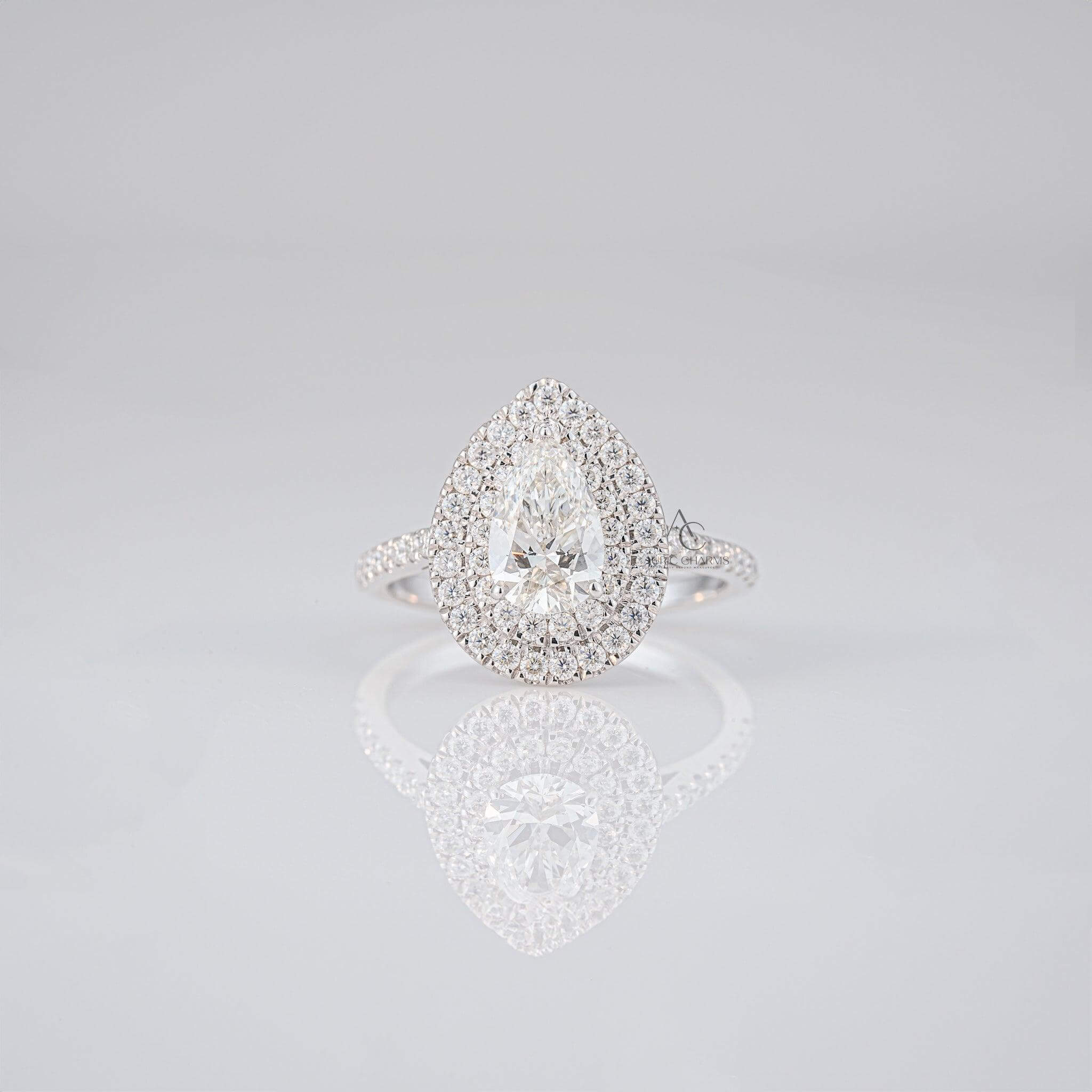Pear-shaped diamond ring with a double halo of diamonds on a reflective surface.