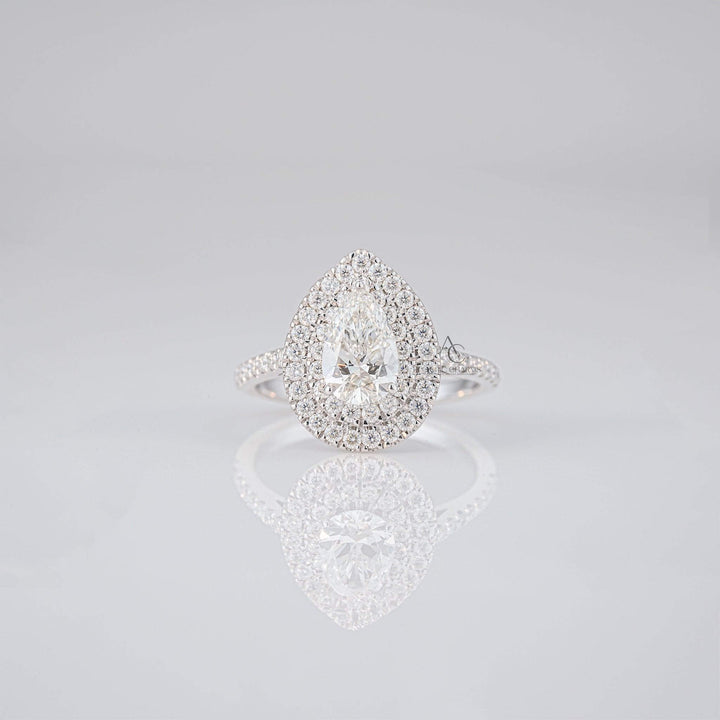 Pear-shaped diamond ring with a double halo of diamonds on a reflective surface.